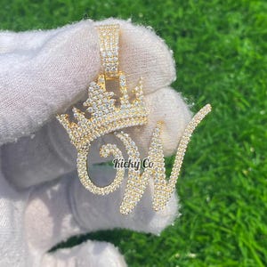 Könnte beinhalten: Goldfarbener Anhänger mit einem Kronen-Design über dem Buchstaben "W", besetzt mit glitzernden Steinen. Der Anhänger enthält den Text "Kicky Co". Der Schmuck wird vor einem grünen Hintergrund gehalten.