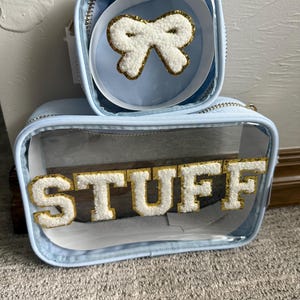 Op de afbeelding: Twee lichtblauwe cosmetische tassen. De grotere tas heeft het woord "STUFF" in witte, donzige letters met gouden rand. De kleinere tas bevat een witte strik met gouden rand. Beide tassen hebben doorzichtige zijkanten en gouden ritsen.