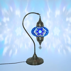 May include: A decorative table lamp with a blue mosaic glass shade featuring a star design. The lamp has a dark metal base and a curved arm. The shade is made of small, colorful glass pieces. The lamp is plugged in.