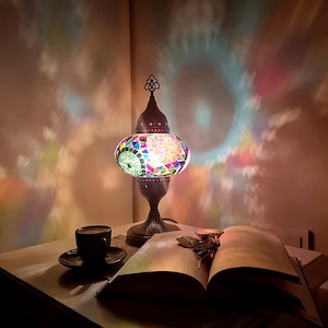 May include: A colorful mosaic table lamp with a dark metal base and a decorative top. The lamp is lit, casting a rainbow of colors on the wall. A cup of coffee and an open book are on the table.