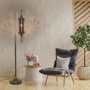 May include: An ornate floor lamp with a dark metal shade and base, casting a patterned light. A grey armchair with a white cushion is next to a small side table with a clock, flowers, and a figurine. The room has a neutral colour scheme.
