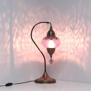 May include: A decorative table lamp with a pink glass shade and a bronze-colored metal base and curved arm. The shade has a textured, mosaic-like appearance. The lamp is turned on, casting a warm glow and patterned shadows on the wall.