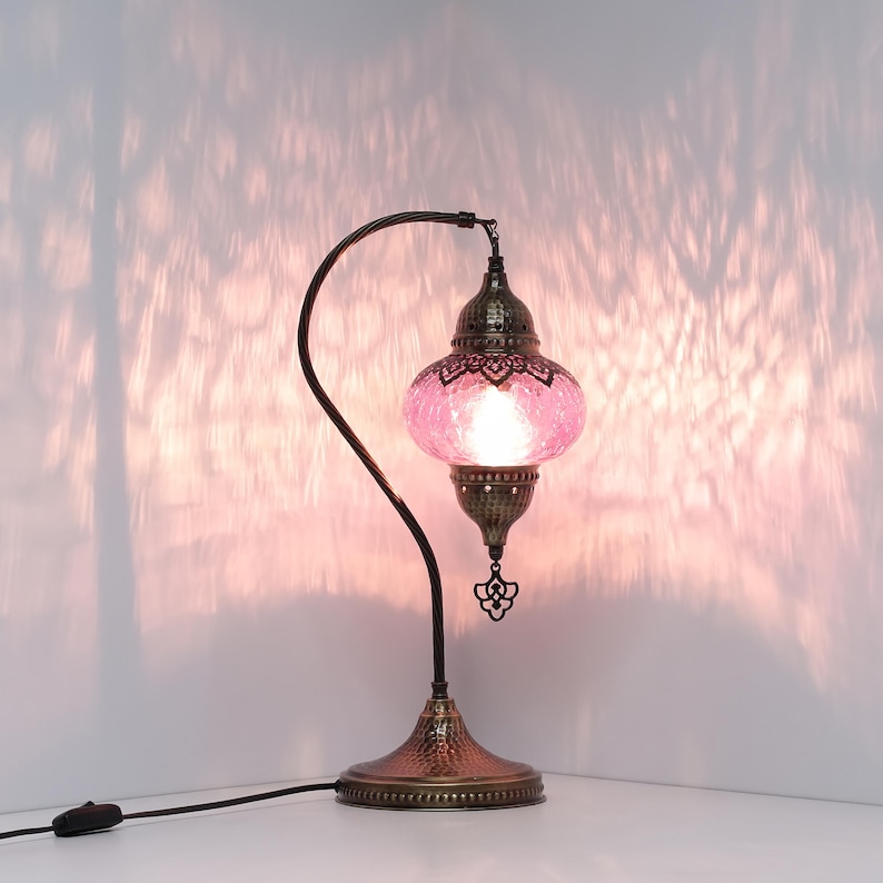 May include: A decorative table lamp with a pink glass shade and a bronze-colored metal base and curved arm. The shade has a textured, mosaic-like appearance. The lamp is turned on, casting a warm glow and patterned shadows on the wall.