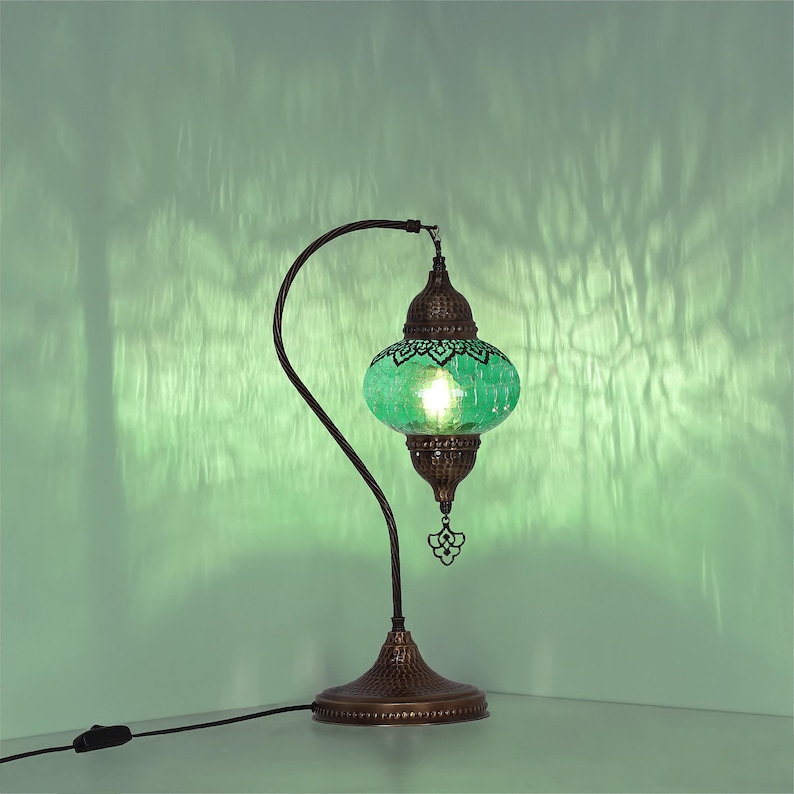 May include: A decorative table lamp with a green glass shade and a bronze-colored metal base and curved arm. The shade has a mosaic design, and the lamp is plugged into an electrical outlet.