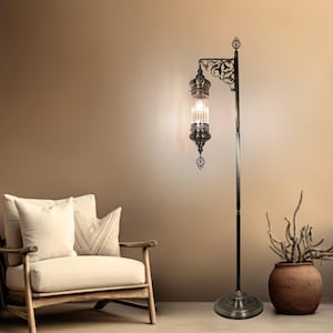 May include: A bronze-colored floor lamp with an ornate, cylindrical glass shade. The lamp features decorative filigree and a round base. The lamp is next to a wooden armchair with cream-colored cushions and a terracotta pot with dried plants.