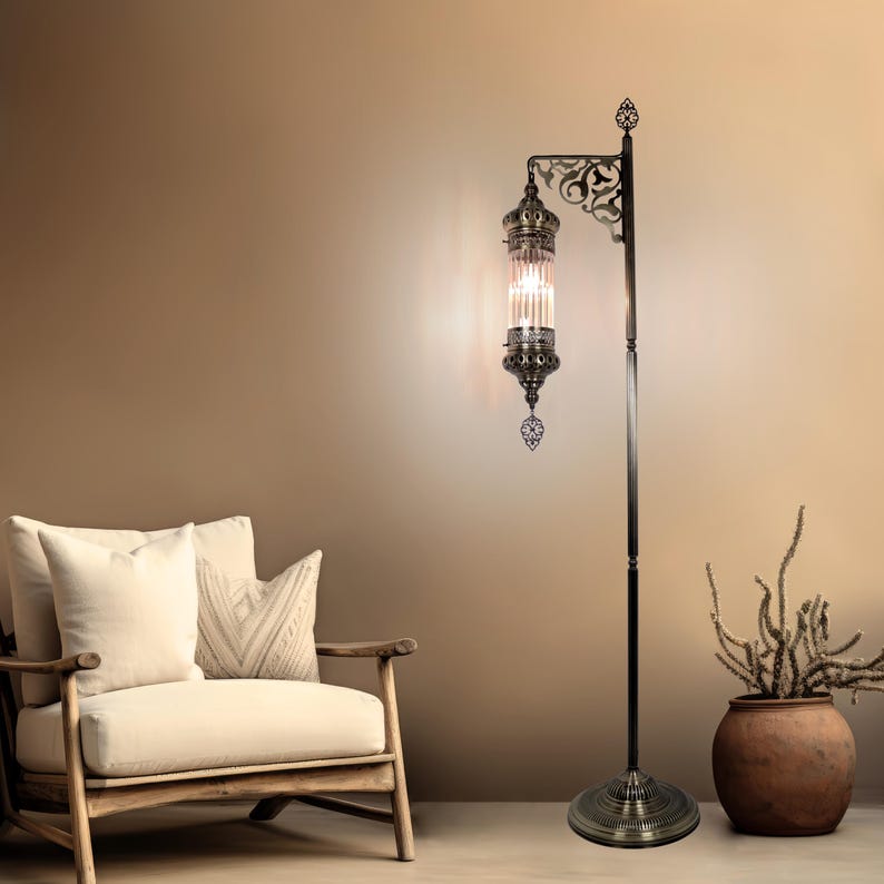 May include: A bronze-colored floor lamp with an ornate, cylindrical glass shade. The lamp features decorative filigree and a round base. The lamp is next to a wooden armchair with cream-colored cushions and a terracotta pot with dried plants.