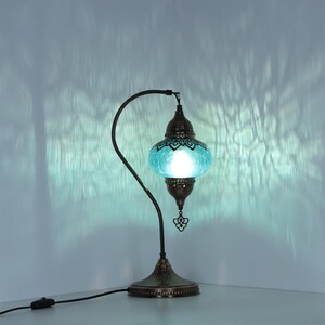 May include: A decorative table lamp with a teal glass shade and a bronze-colored metal base and curved arm. The lamp has an ornate design and is lit, casting a soft glow. The lamp is plugged into an electrical outlet.