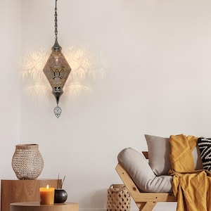 Puede incluir: Una lámpara colgante ornamental en forma de diamante con intrincados patrones calados, que proyecta patrones de luz decorativos en la pared. La lámpara está suspendida de una cadena y presenta un remate decorativo. El acabado metálico de la lámpara es oscuro.