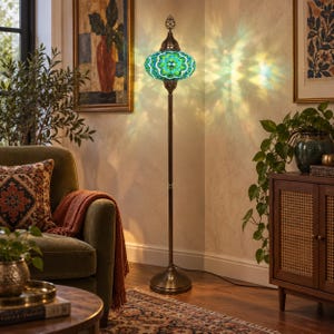 May include: A tall floor lamp featuring a mosaic glass shade in shades of teal, blue, and white. The lamp has a slender, dark metal stand with a decorative finial at the top and a wide, circular base, casting a colorful light pattern on the wall.
