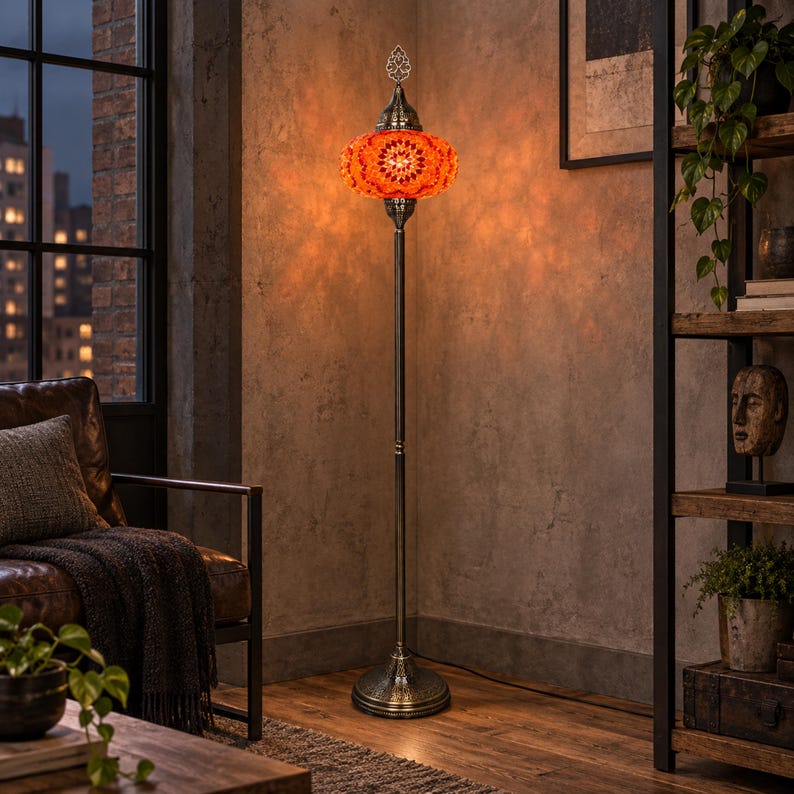 May include: A tall floor lamp featuring a vibrant orange mosaic glass shade with intricate patterns. The lamp has a slender, dark metallic stand and a circular base with ornate detailing. The light emits a warm, glowing orange hue.