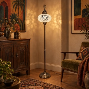 May include: A tall floor lamp featuring a round, white mosaic glass shade with a dark metal base and a decorative finial. The lamp emits a warm, glowing light that casts intricate, patterned shadows on the surrounding wall and floor surfaces.