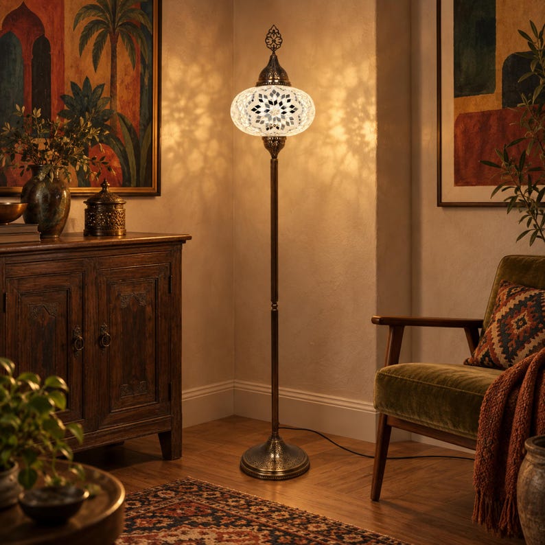 May include: A tall floor lamp featuring a round, white mosaic glass shade with a dark metal base and a decorative finial. The lamp emits a warm, glowing light that casts intricate, patterned shadows on the surrounding wall and floor surfaces.