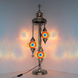 May include: A decorative table lamp with a bronze finish and three hanging mosaic glass shades. The shades feature a colourful floral design with red, yellow, blue, and green hues. The lamp has a round base and a tall, ornate stem.