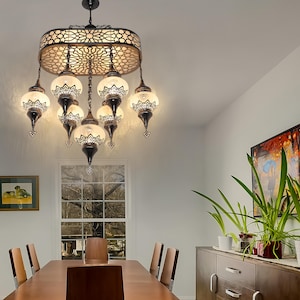 May include: An ornate chandelier with a bronze-colored frame and eight glass globes. The frame features a decorative, openwork design. The glass globes have a white and black floral pattern. The chandelier hangs above a wooden dining table.