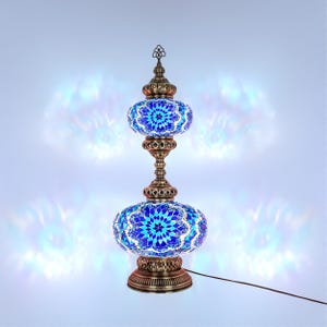 Peut inclure: Une lampe de table décorative avec un motif en mosaïque. La lampe présente deux abat-jours ovales et un pied et une tige en métal de couleur bronze. Les abat-jours sont ornés de morceaux de verre bleus et blancs disposés en motif floral. Un cordon d'alimentation part de la base.