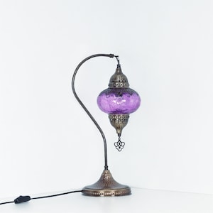 May include: A decorative table lamp with a purple glass shade and a bronze-colored metal base and curved arm. The lamp has an ornate design and a textured finish, with a power cord and on/off switch.