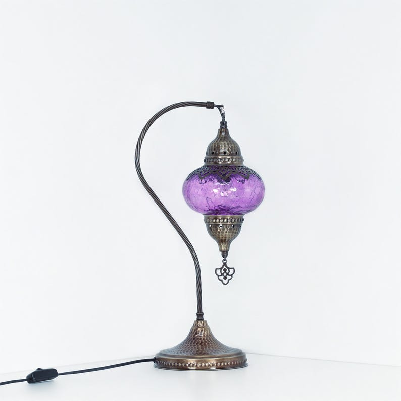 May include: A decorative table lamp with a purple glass shade and a bronze-colored metal base and curved arm. The lamp has an ornate design and a textured finish, with a power cord and on/off switch.