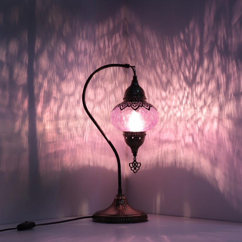 May include: A decorative table lamp with a pink glass shade and a dark metal frame. The lamp has an ornate design with a curved arm and a detailed base. The light casts a patterned shadow on the wall.