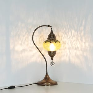 May include: A decorative table lamp with a yellow, patterned glass shade and a bronze-colored metal base and curved arm. The lamp has an ornate design and a small decorative charm hanging from the bottom.