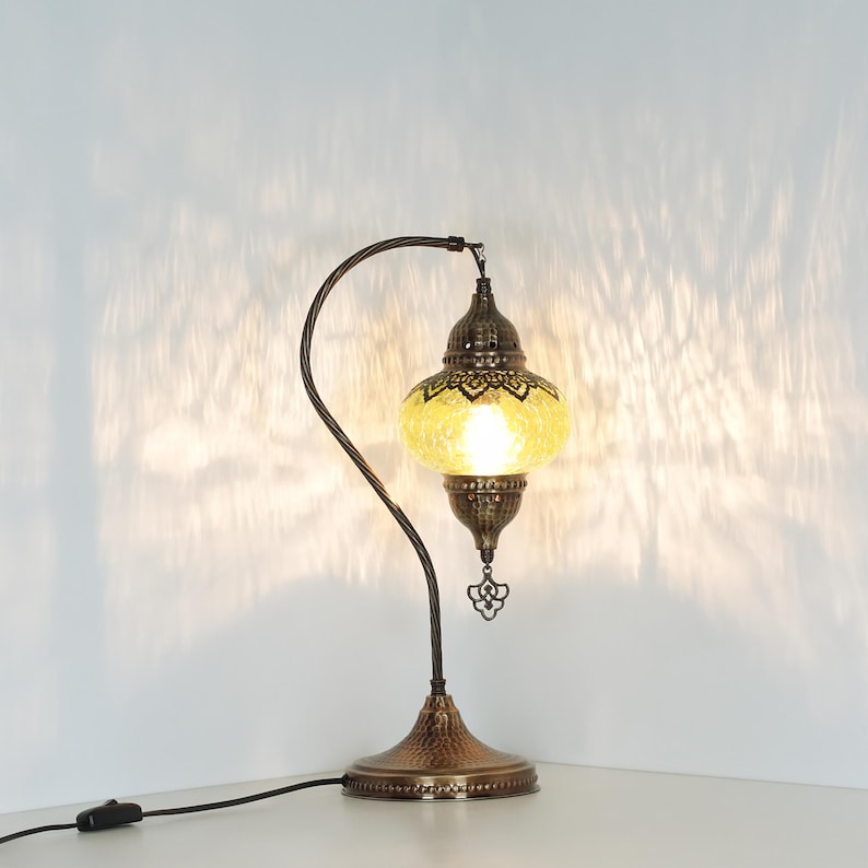 May include: A decorative table lamp with a yellow, patterned glass shade and a bronze-colored metal base and curved arm. The lamp has an ornate design and a small decorative charm hanging from the bottom.