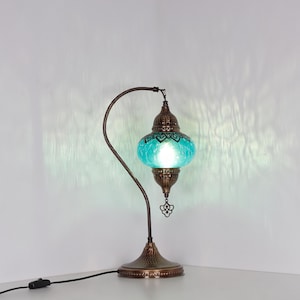 May include: A decorative table lamp with a turquoise glass shade and a bronze-colored metal base and curved arm. The lamp has an ornate design and a small decorative pendant hanging from the bottom.