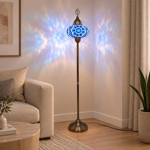 May include: A tall floor lamp with a bronze-colored metal stand and a round base. The lamp features a spherical shade crafted from blue and white mosaic glass pieces, casting intricate, star-shaped light patterns onto the surrounding walls.