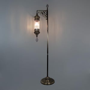 May include: An antique-style floor lamp with a bronze finish. The lamp features a decorative, cylindrical glass shade with vertical lines, suspended from a curved arm. The base is round and ornate, and the lamp stands approximately 60 inches tall.