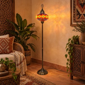 May include: A tall floor lamp featuring a vibrant, multi-colored mosaic glass shade with geometric patterns in orange, yellow, and blue. The lamp has a slender, dark metal stand and a circular base, emitting a warm, glowing light onto the wall.