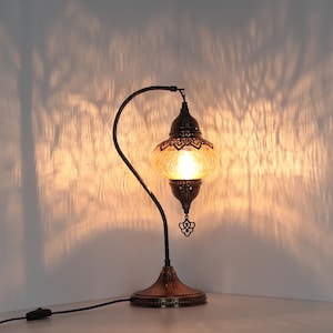 May include: A decorative table lamp with a curved, dark metal frame and a textured, amber-colored glass shade. The lamp has an ornate, bronze-colored base and a dangling decorative element. The light casts a warm glow and interesting shadows on the wall.
