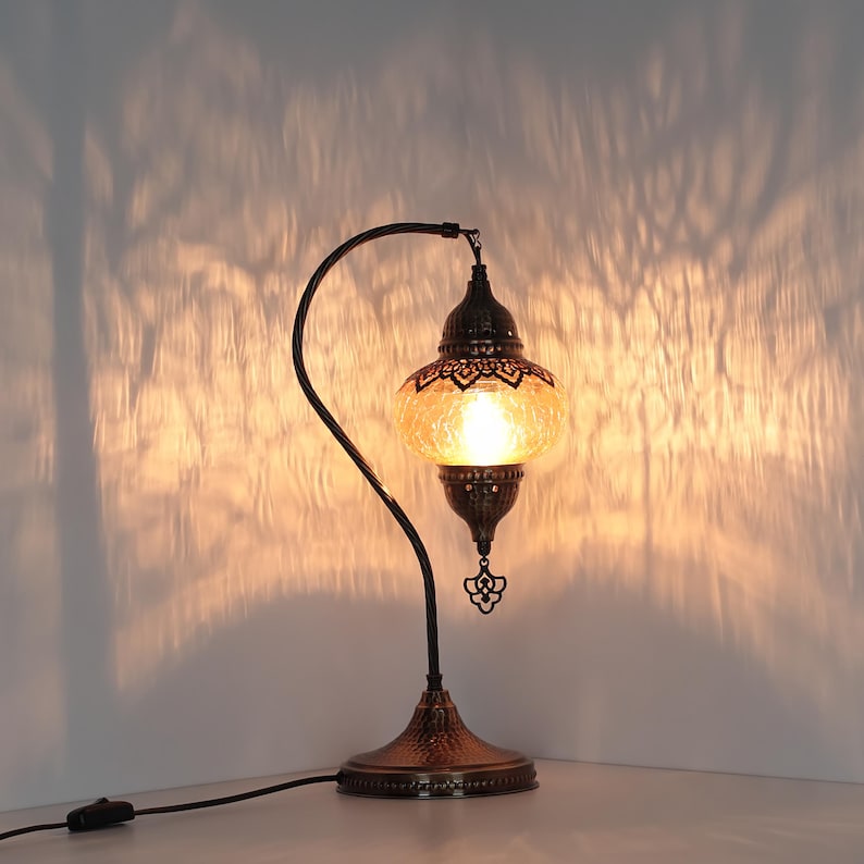 May include: A decorative table lamp with a curved, dark metal frame and a textured, amber-colored glass shade. The lamp has an ornate, bronze-colored base and a dangling decorative element. The light casts a warm glow and interesting shadows on the wall.