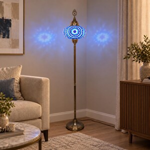 May include: A tall floor lamp featuring a brass-toned metal stand and a round, mosaic glass shade. The shade displays a geometric pattern in shades of blue and white, emitting a bright blue light that casts star-shaped patterns on the wall.