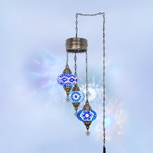 Op de afbeelding: Een hanglamp met drie blauwe mozaïek glazen bollen. De bollen hangen aan een decoratieve metalen armatuur en ketting. Het mozaïekglas heeft een mix van blauwe en witte patronen. Geschikt voor interieurdecoratie.