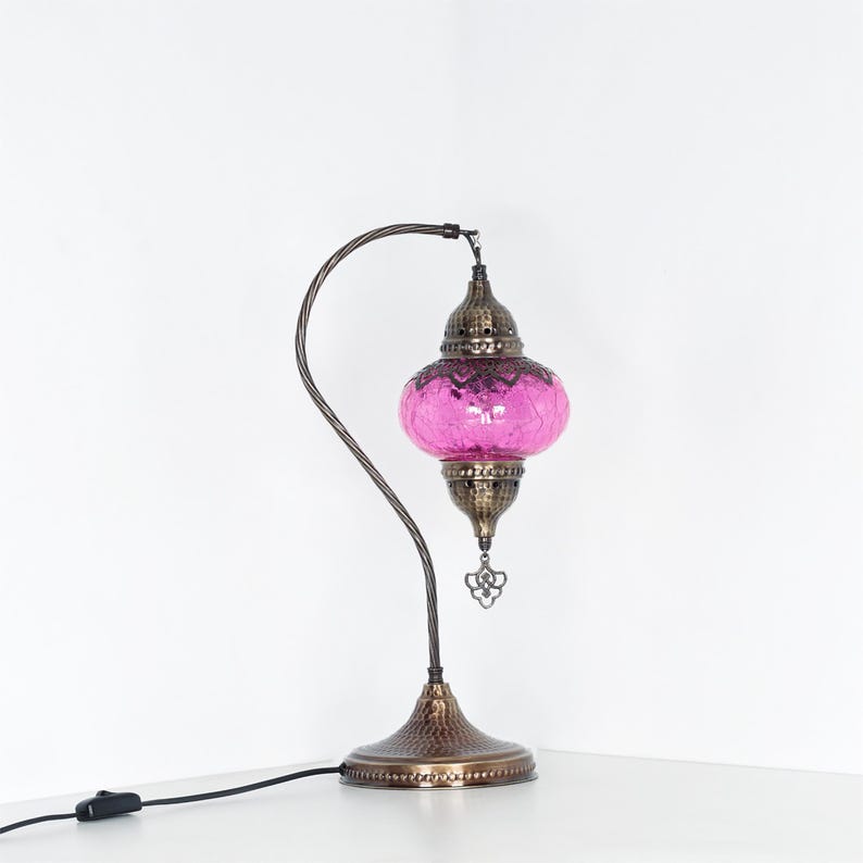 May include: A decorative table lamp with a curved, bronze-colored metal frame. The lamp features a pink, glass shade with a mosaic pattern. The base and top of the lamp are also bronze-colored, with intricate detailing. The lamp is plugged into a wall outlet.