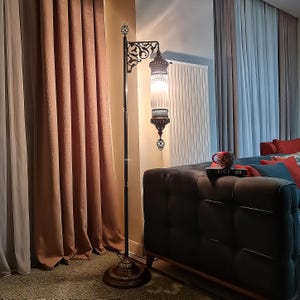 May include: A tall, ornate floor lamp with a dark metal frame and a decorative, glass lantern-style shade. The lamp features intricate filigree details and a warm, inviting glow, perfect for adding ambiance to a living space. The lamp is approximately 6 feet tall.