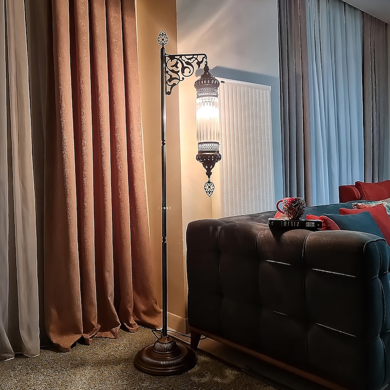 May include: A tall, ornate floor lamp with a dark metal frame and a decorative, glass lantern-style shade. The lamp features intricate filigree details and a warm, inviting glow, perfect for adding ambiance to a living space. The lamp is approximately 6 feet tall.