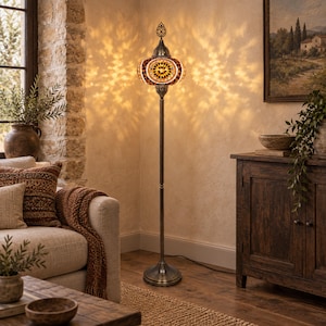 May include: A tall floor lamp featuring a mosaic glass shade with amber, brown, and white geometric patterns. The lamp has a slender, dark metal stand and a circular base, casting intricate light patterns on the wall behind it in a room.