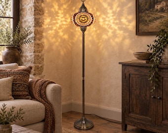 May include: A tall floor lamp featuring a mosaic glass shade with amber, brown, and white geometric patterns. The lamp has a slender, dark metal stand and a circular base, casting intricate light patterns on the wall behind it in a room.