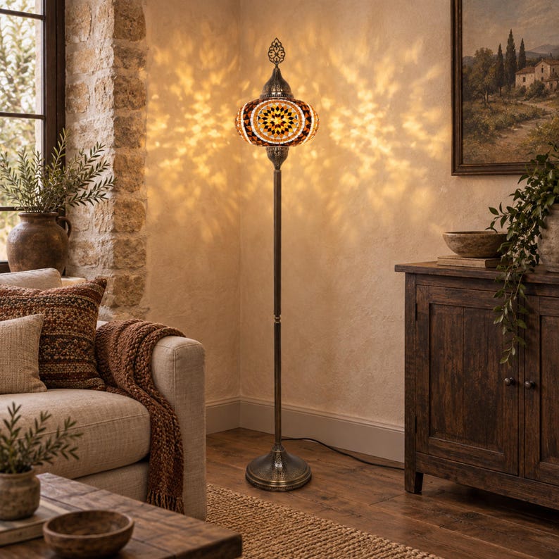 May include: A tall floor lamp featuring a mosaic glass shade with amber, brown, and white geometric patterns. The lamp has a slender, dark metal stand and a circular base, casting intricate light patterns on the wall behind it in a room.
