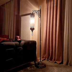 May include: A tall, ornate floor lamp with a dark brown metal frame and a decorative, patterned shade. The lamp has a round base and a curved arm holding the glass shade. The shade is lit, casting a warm glow in the room. The lamp is next to a brown couch and pink curtains.