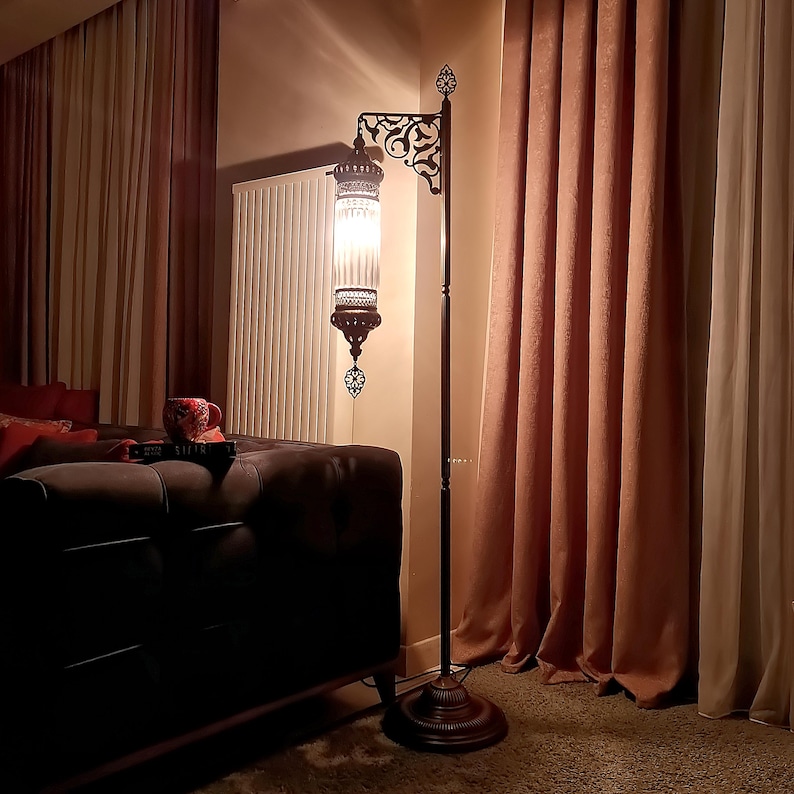 May include: A tall, ornate floor lamp with a dark brown metal frame and a decorative, patterned shade. The lamp has a round base and a curved arm holding the glass shade. The shade is lit, casting a warm glow in the room. The lamp is next to a brown couch and pink curtains.