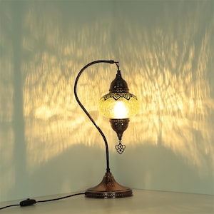 May include: A decorative table lamp with a mosaic glass shade in shades of yellow and brown. The lamp has a dark bronze-colored metal base and a curved arm. The lamp is plugged into an electrical outlet.