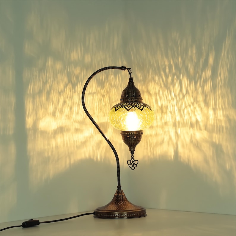 May include: A decorative table lamp with a mosaic glass shade in shades of yellow and brown. The lamp has a dark bronze-colored metal base and a curved arm. The lamp is plugged into an electrical outlet.
