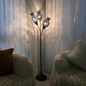 May include: A decorative floor lamp with three flower-shaped lampshades. The lampshades have a black and white pattern and are lit from within. The lamp has a dark metal stem and base, and is placed in front of white curtains.