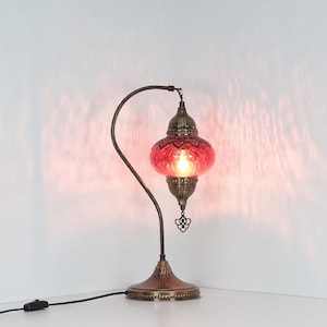May include: A decorative table lamp with a red glass shade and a bronze-colored metal base and curved arm. The shade has a textured surface and a decorative pattern. The lamp is plugged into an electrical outlet.