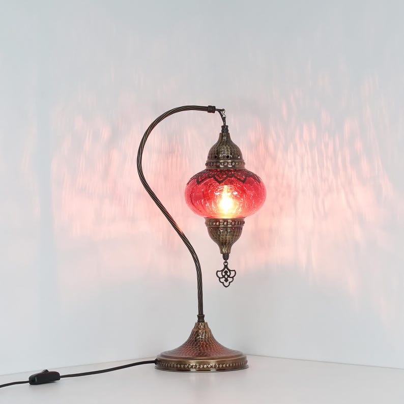 May include: A decorative table lamp with a red glass shade and a bronze-colored metal base and curved arm. The shade has a textured surface and a decorative pattern. The lamp is plugged into an electrical outlet.