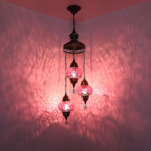 Può includere: Una lampada a sospensione decorativa con tre globi di vetro rosa. La lampada ha una cornice e una catena in metallo bronzato ornate. La luce proietta un bagliore rosa sulle pareti circostanti, creando un'atmosfera calda. La lampada è progettata per uso interno.