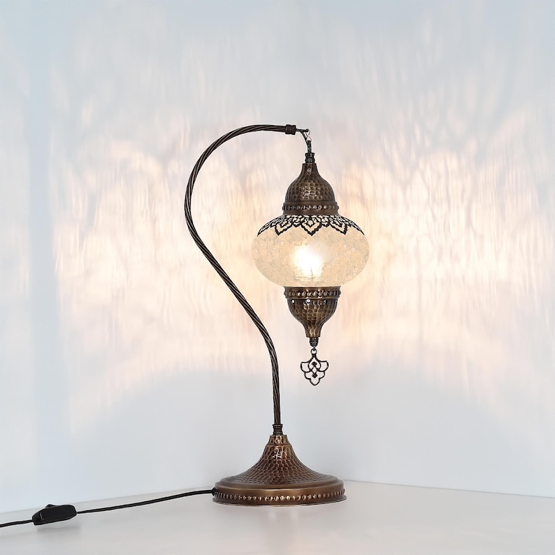 May include: A decorative table lamp with a bronze-colored base and curved arm. The lamp features a globe-shaped shade with a mosaic pattern and intricate metalwork. The lamp is plugged into a wall outlet.