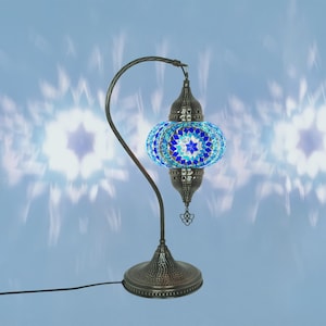 May include: A decorative table lamp with a mosaic glass shade in shades of blue and white. The lamp has a curved, dark metal stand and a round base. The shade is a globe shape with a starburst pattern. The lamp is plugged into an electrical outlet.