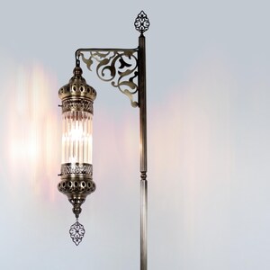 May include: An antique-style floor lamp with a bronze finish. The lamp features a decorative, ornate design with a clear, cylindrical glass shade. The lamp has a detailed, filigree-style bracket and a decorative finial at the top.