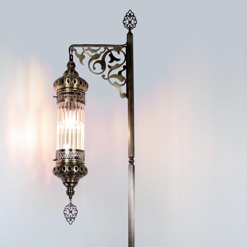 May include: An antique-style floor lamp with a bronze finish. The lamp features a decorative, ornate design with a clear, cylindrical glass shade. The lamp has a detailed, filigree-style bracket and a decorative finial at the top.